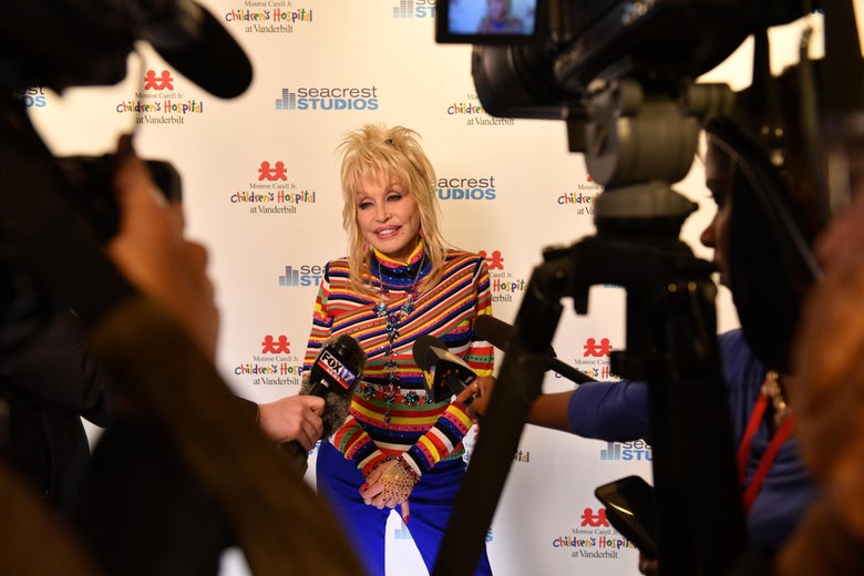 Wearing a red, orange, blue, green and white striped shirt, Dolly Parton poses with film cameras, tripods and microphones in the foreground